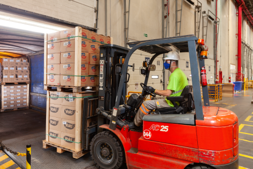 A worker in a hard hat operates a red forklift to load boxes onto a truck in a warehouse setting.