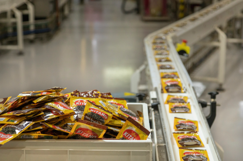 Factory conveyor belt with packaged products moving alongside a bin filled with similar packets.