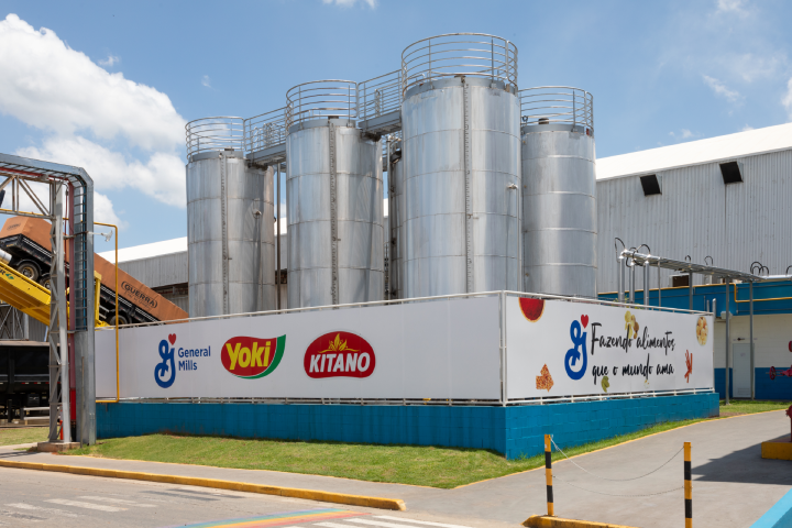 Industrial facility with several large silver silos with a General mills, Yoki and Kitano Brand logos on its surface, standing tall in a rural landscape against a clear sky.