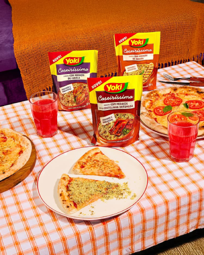 Two pizzas and slices on plates with breaded seasoning packets and glasses of red drinks on a checkered tablecloth.