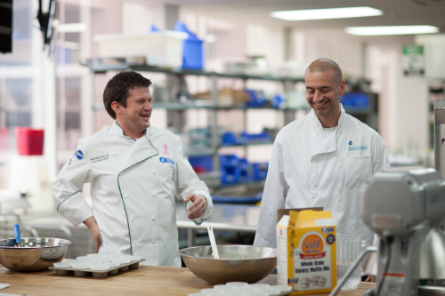 Two chefs in a commercial kitchen are standing near a table with mixing bowls and baking ingredients.